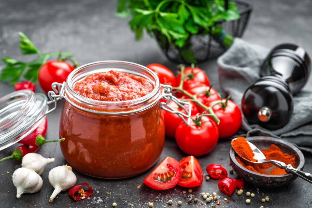 L'image montre un bocal de sauce tomate maison, entouré de divers ingrédients utilisés pour la préparation. On peut voir des tomates fraîches, des gousses d'ail, des piments rouges, des épices moulues, et des herbes vertes en arrière-plan. Le bocal est en verre avec un couvercle en métal et est rempli de sauce tomate rouge et épaisse.