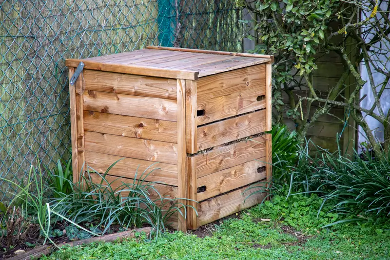 Un bac à compost en bois fermé, placé dans un coin de jardin entouré de végétation et d’une clôture.

