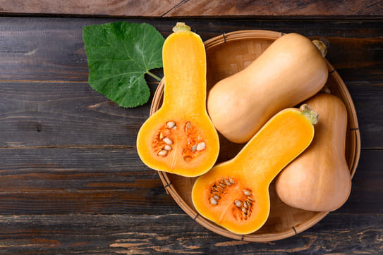 une courge butternut coupée en deux, révélant l'intérieur orange vif et les graines. À côté, on voit aussi des butternuts entières posées dans un panier. Cette vue est typique pour identifier la maturité intérieure du fruit.